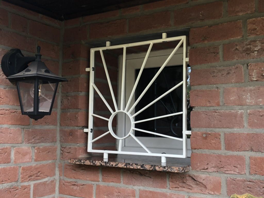 Weiß beschichtetes Fenstergitter mit Sonnenmotiv an einer Klinkerfassade.