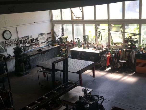 Werkstatt von oben mit Lochraster-Schweißtisch, Werkbänken und Maschinen vor einer Fensterfront.